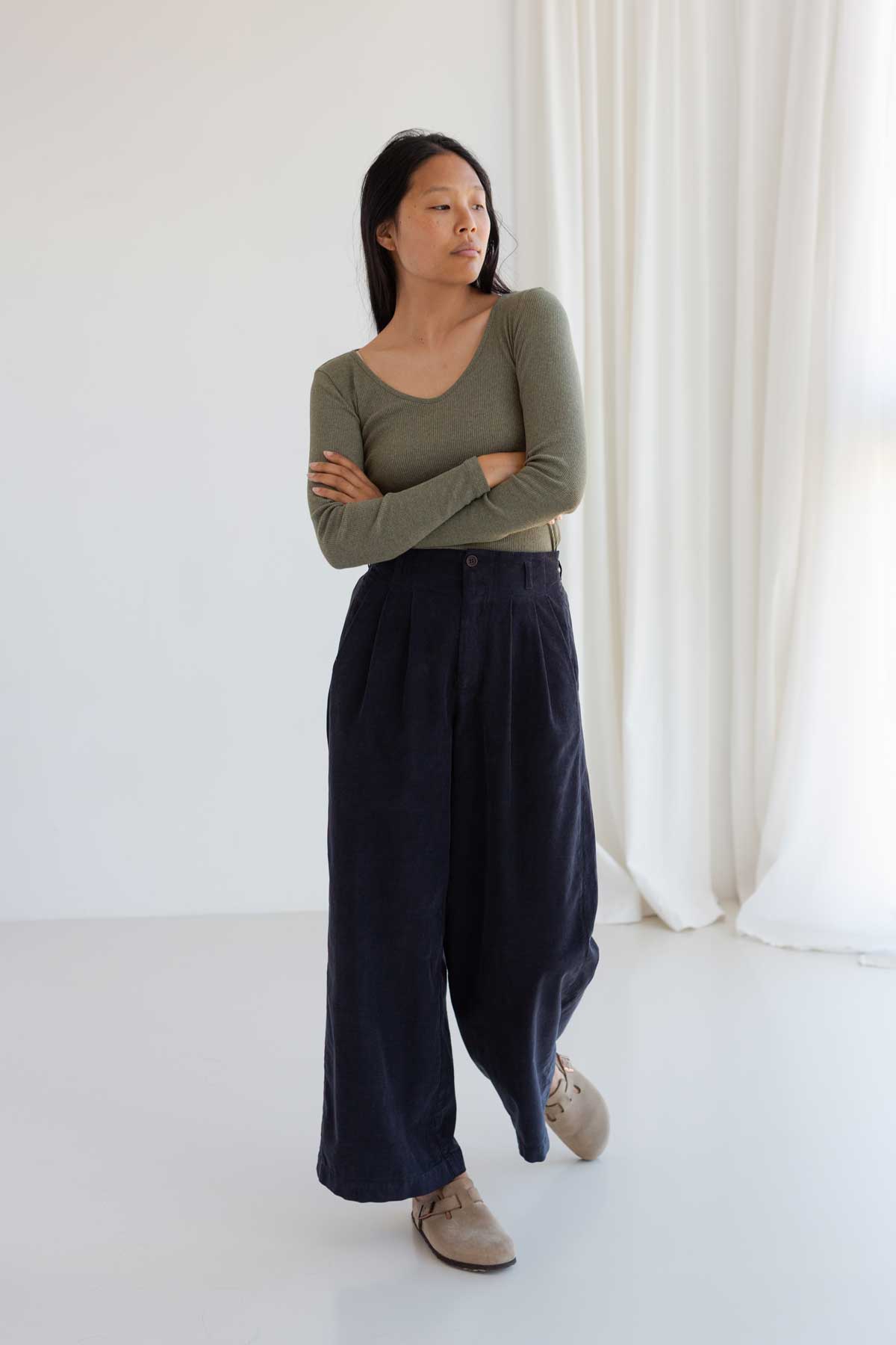 Woman with arms crossed in olive green scoop-neck ribbed top and wide-leg dark blue corduroy pants.
